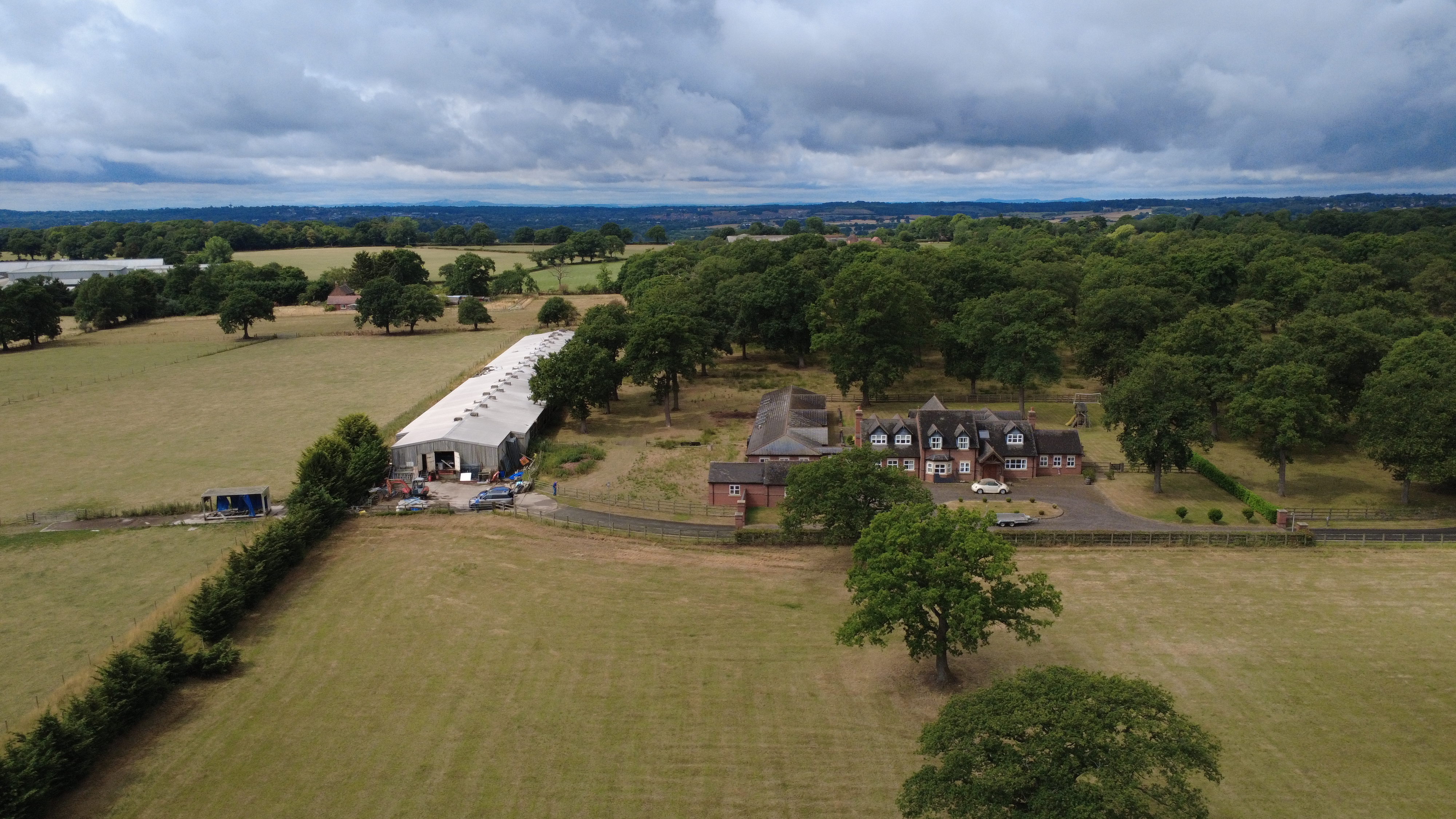 Aerial view of a rural property featuring a large house surrounded by greenery, fields, and a barn under a cloudy sky.